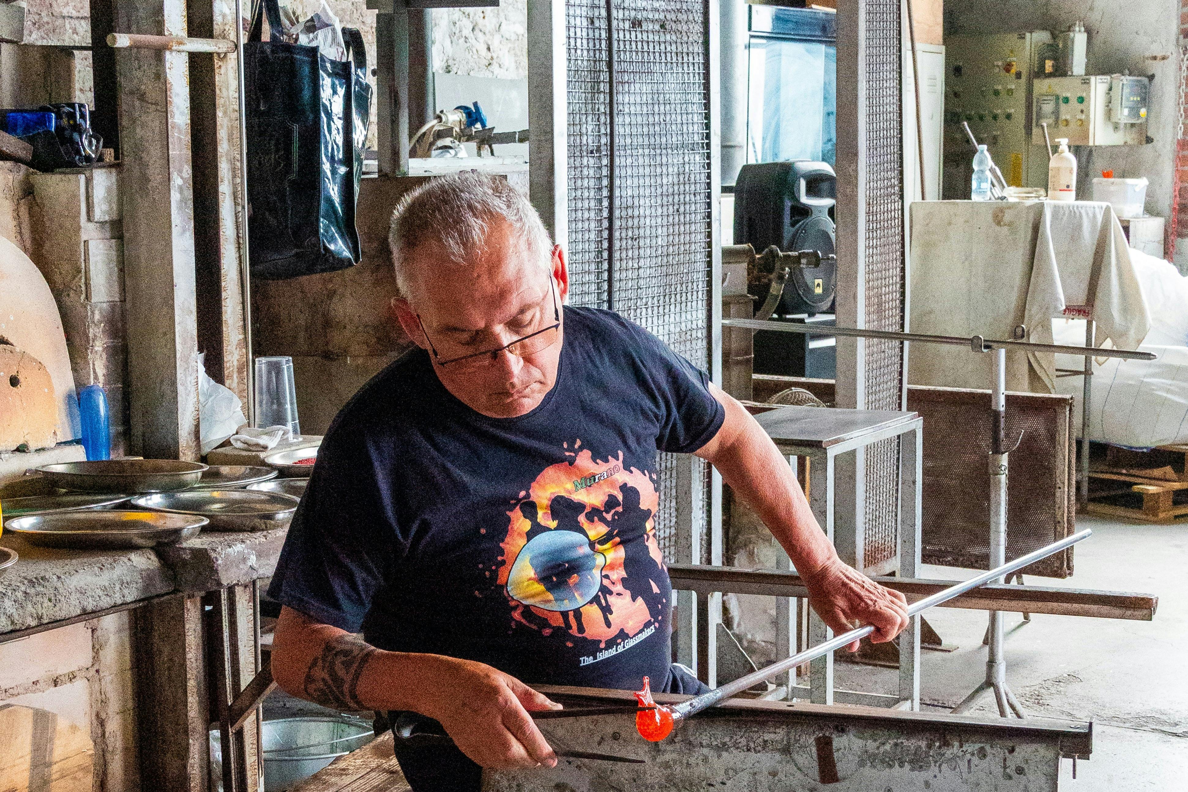 A man works on glassblowing, shaping molten glass on a rod in an industrial workspace. He wears a black T-shirt with a graphic.