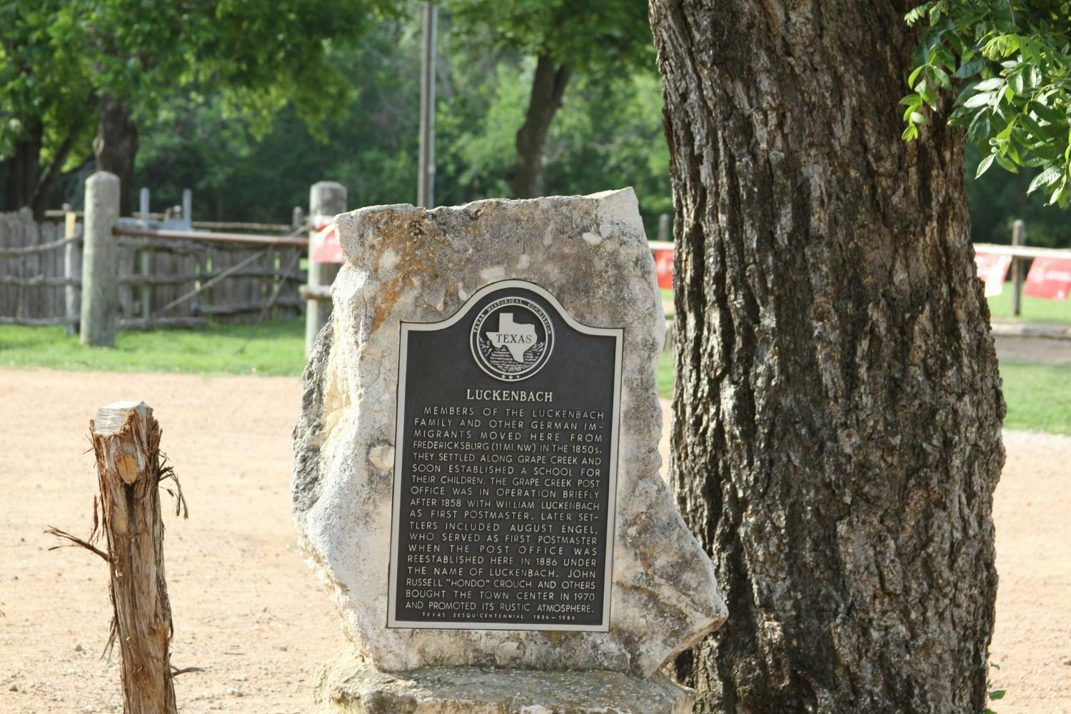 Plaketa umístěná na velkém kameni pod stromem s podrobnostmi o historii Luckenbachu v Texasu. Viditelné zalesněné pozadí.