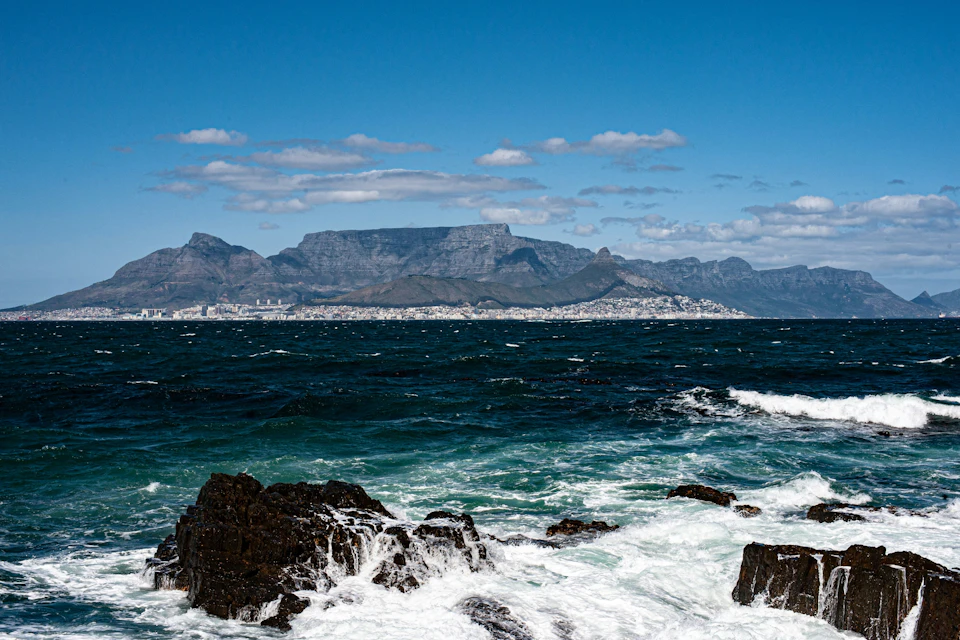 Waves crash against rocks with a distant view of coastal city buildings and a mountain range under a clear blue sky.