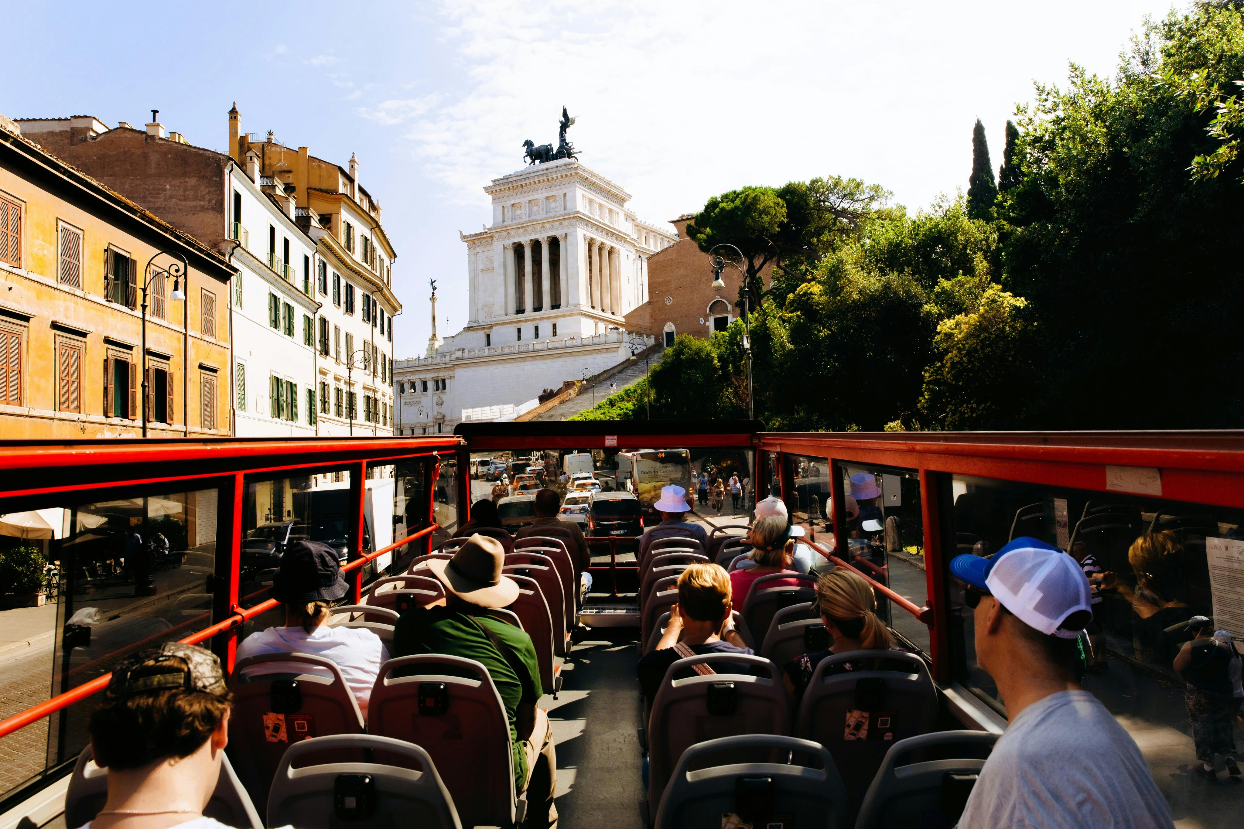 Tourists seated on the upper deck of a red open-top bus passing a historical building with a statue in a sunny urban area.