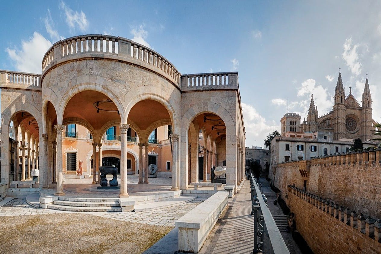 Historická kamenná kolonáda s oblouky před gotickou katedrálou pod modrou oblohou s mraky.