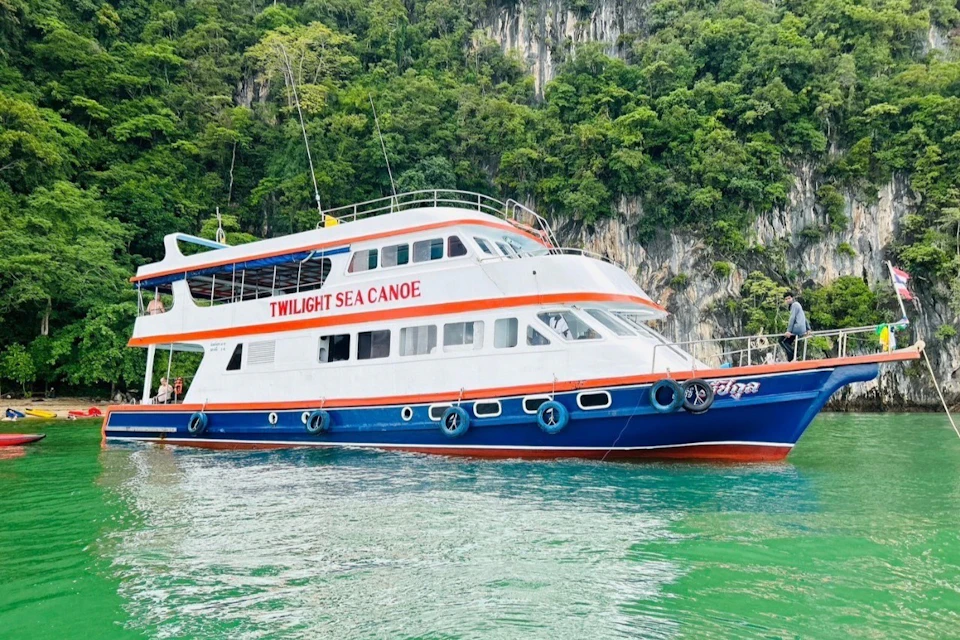 A large boat named "Twilight Sea Canoe" on emerald-green water with a lush, rocky shoreline in the background.