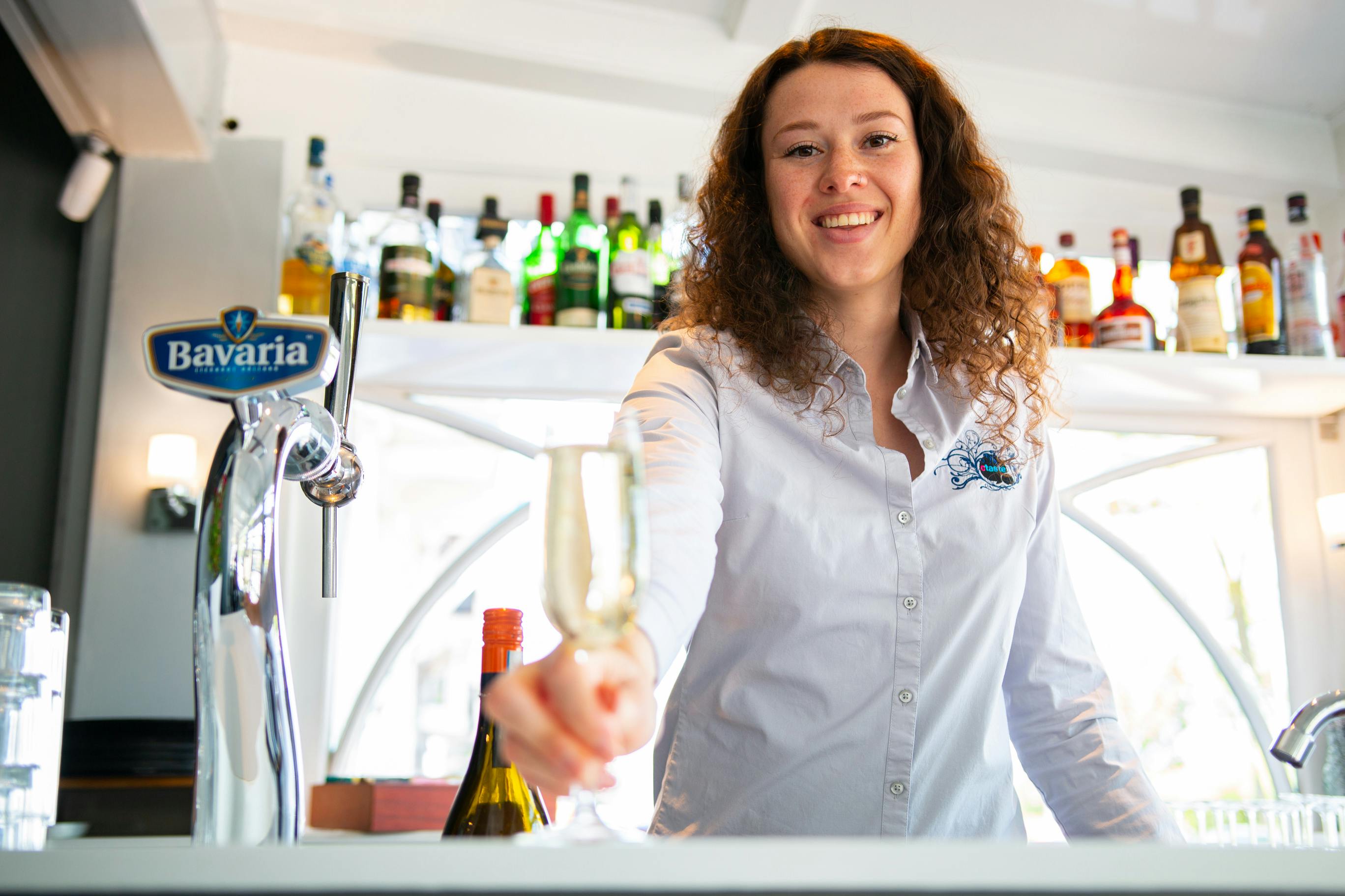 Χαμογελαστός μπάρμαν σε λευκό πουκάμισο κρατώντας ένα ποτήρι, με διάφορα μπουκάλια στα ράφια στο παρασκήνιο.