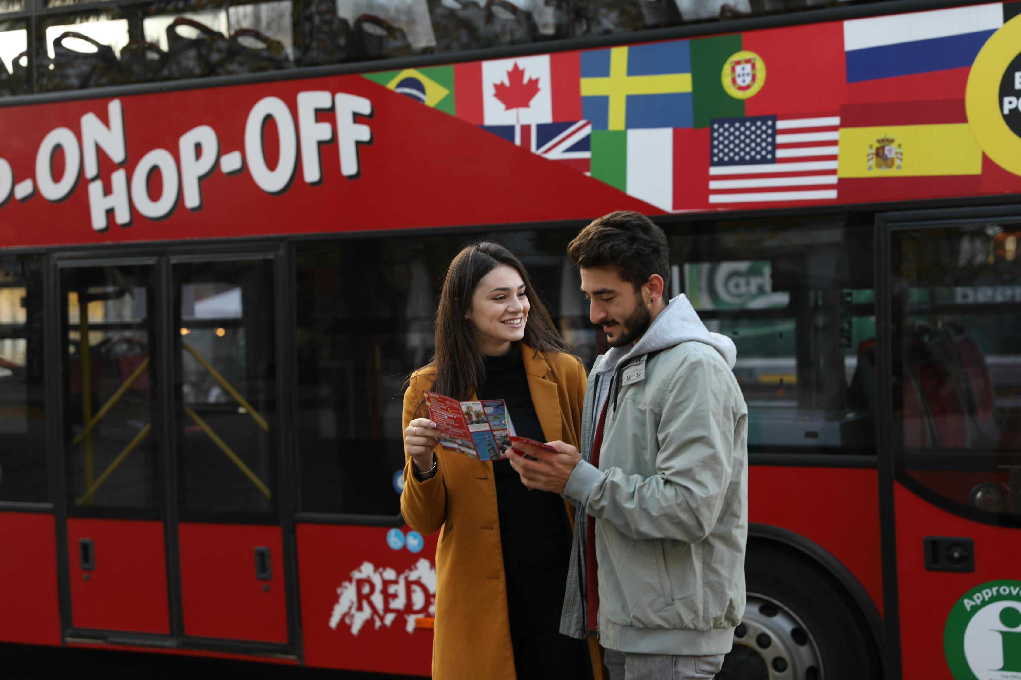 Two people looking at a pamphlet in front of a red hop-on-hop-off bus with various national flags on it.
