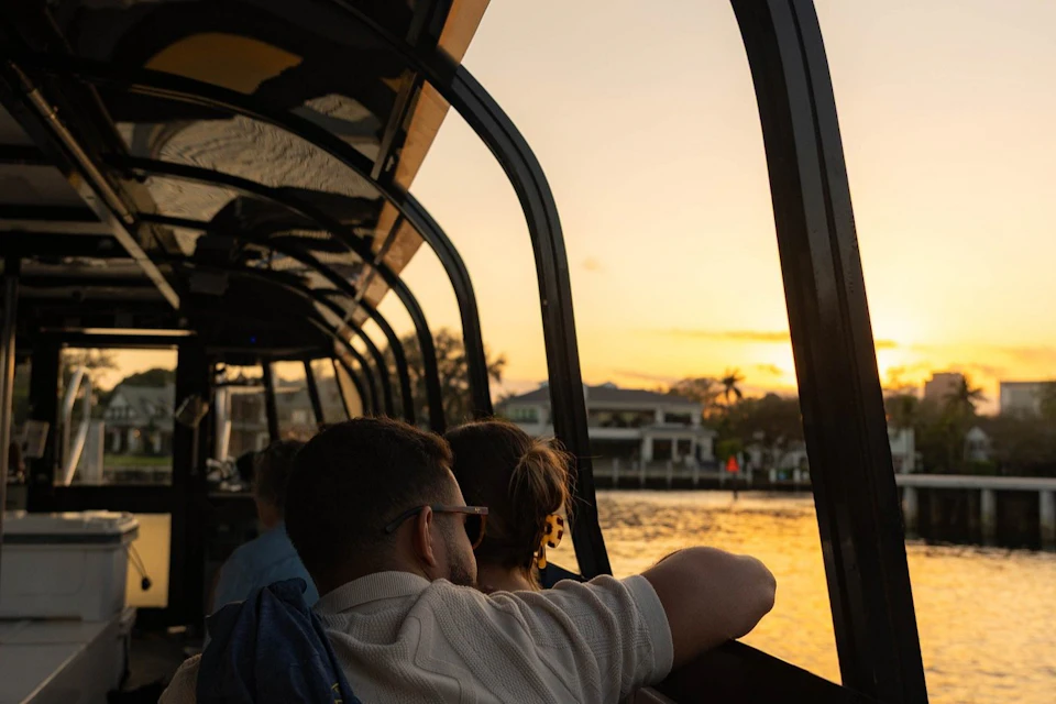 A couple sits on a boat with a glass-enclosed cabin, watching a sunset over a waterfront with buildings and palm trees.