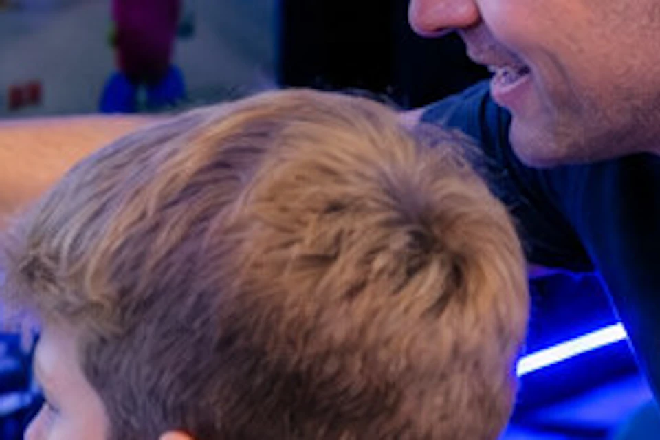 A child in a racing simulator with an adult pointing at a screen in an arcade setting.