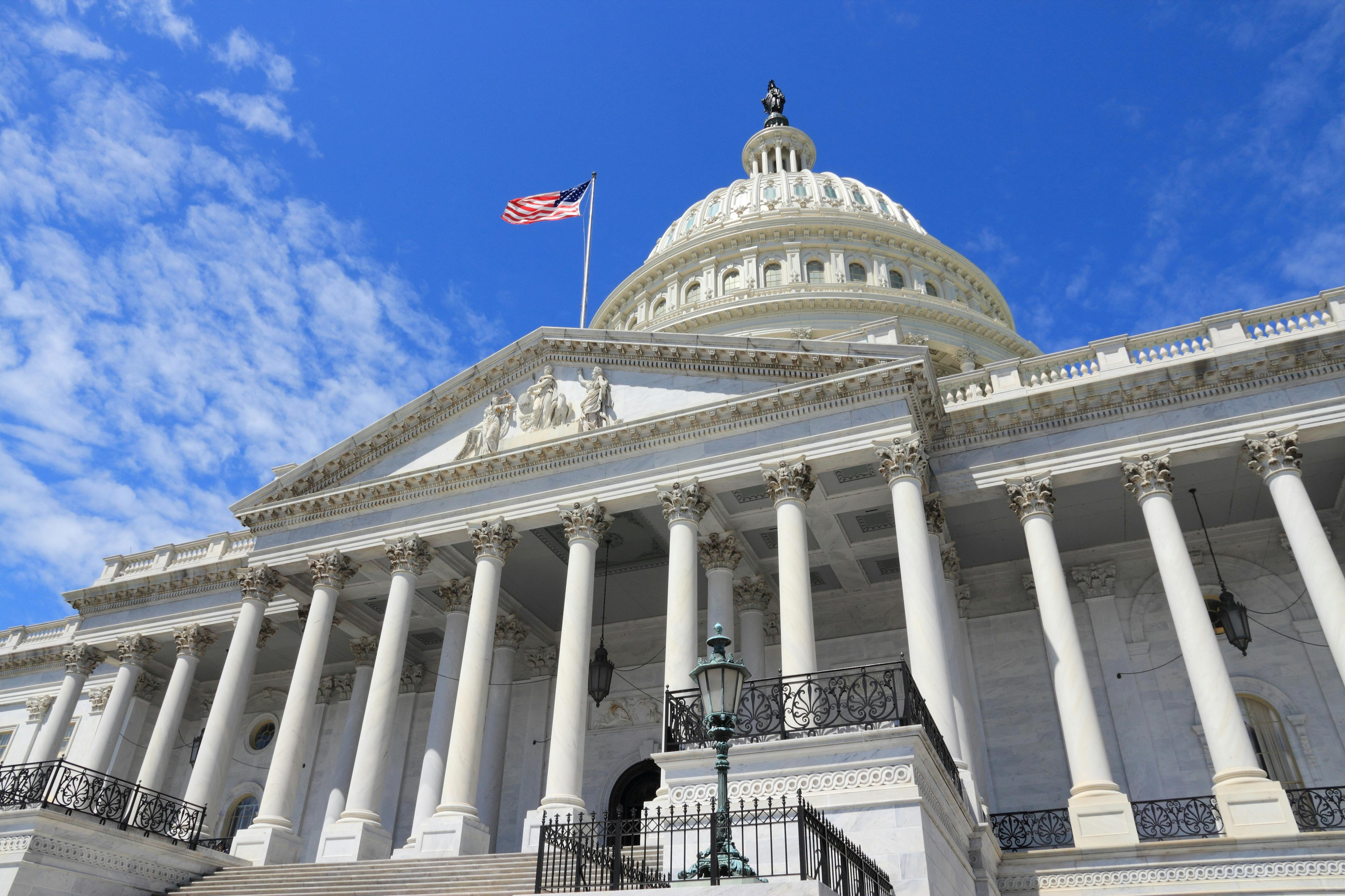 US Capitol-byggnaden med pelare, en amerikansk flagga och en stor kupol under en blå himmel med några moln.