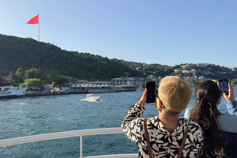 A person on a boat with short blonde hair is taking a photo of a seagull in flight over a body of water near a coastal town.