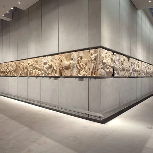 En museiutställning har en omfattande skulptural frise, som visas längs en väl upplyst, minimalistisk hall med grå väggar och taklampor.