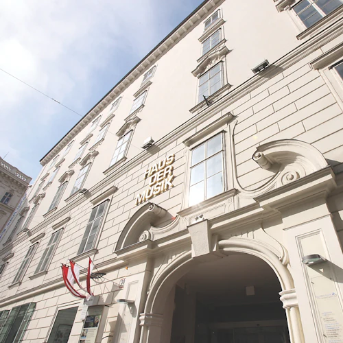 Utsmyckad byggnadsfasad med välvd ingång, stora fönster, "HAUS DER MUSIK" -skylt och rödvit flagga under en solig himmel.