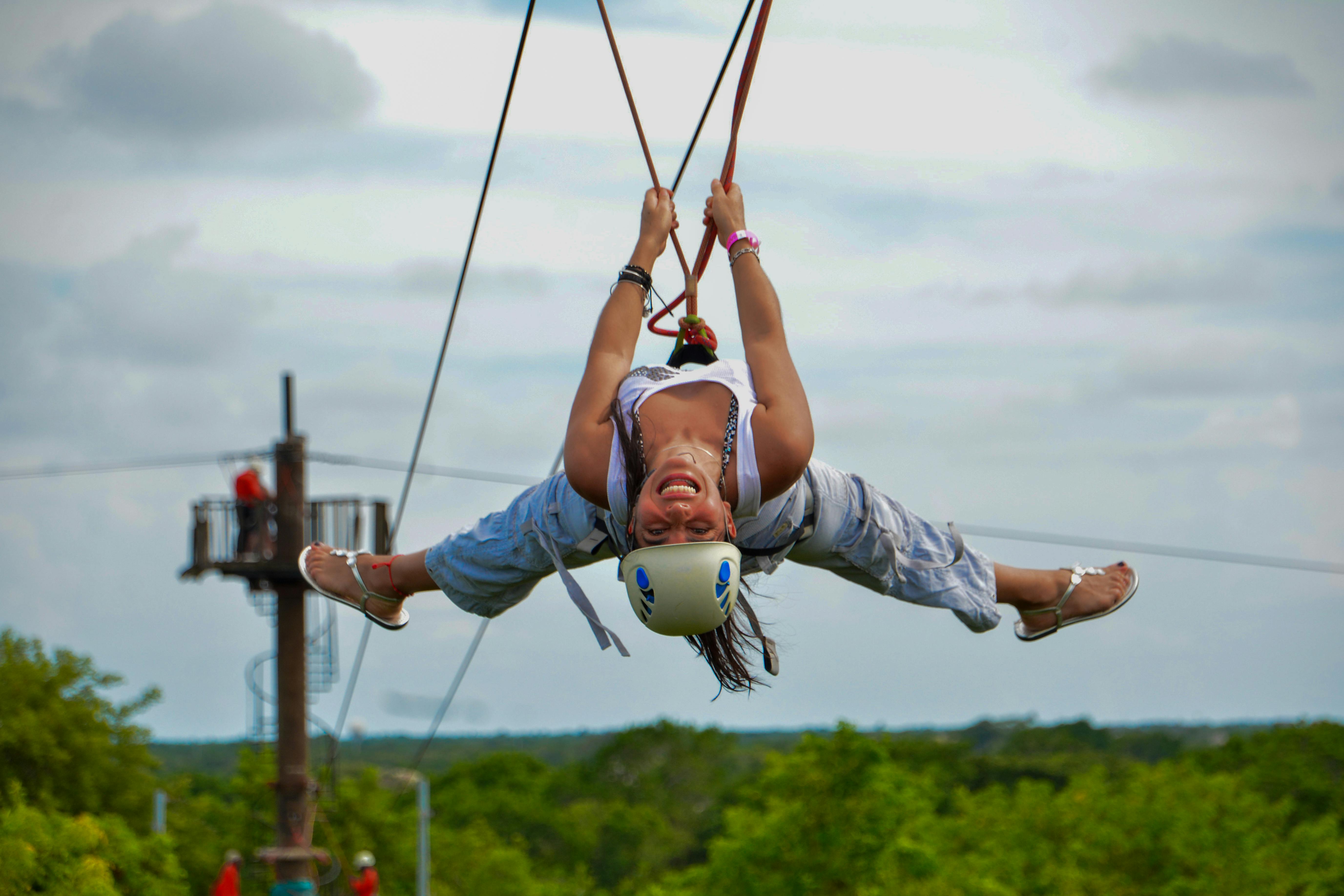 ヘルメットを着てジップラインに逆さまに乗り、笑顔で腕と脚を広げている人。 背景には緑の木。