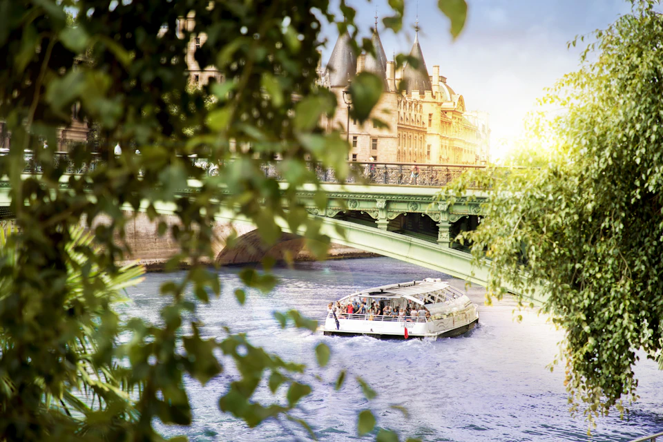 A sightseeing boat sails under a green bridge on a calm river, framed by leafy trees with a historic building in the background.