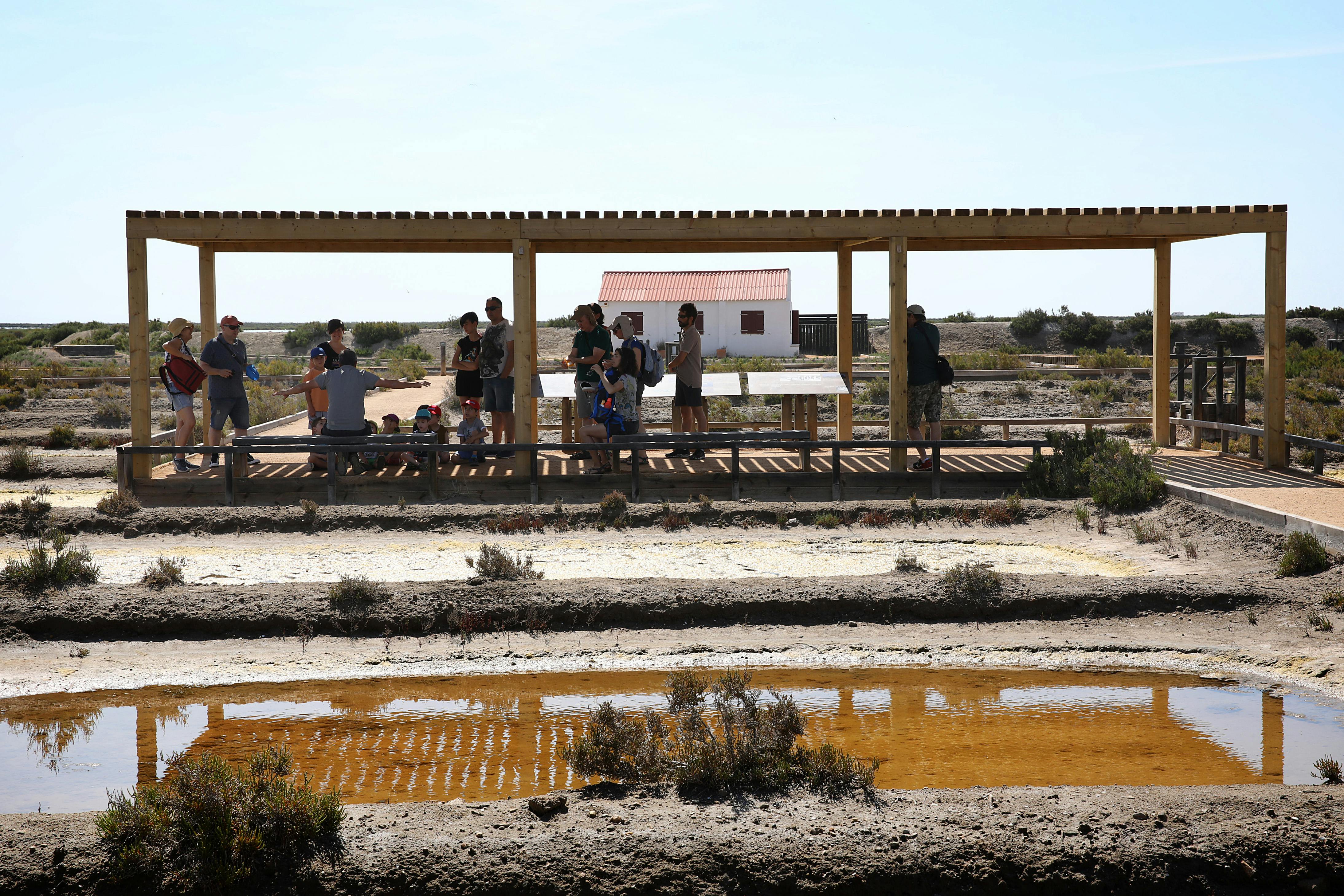 Μια ομάδα ανθρώπων στέκεται κάτω από ένα ανοιχτό ξύλινο καταφύγιο σε μια ξηρή, αμμώδη περιοχή με ρηχά κανάλια νερού και βλάστηση.