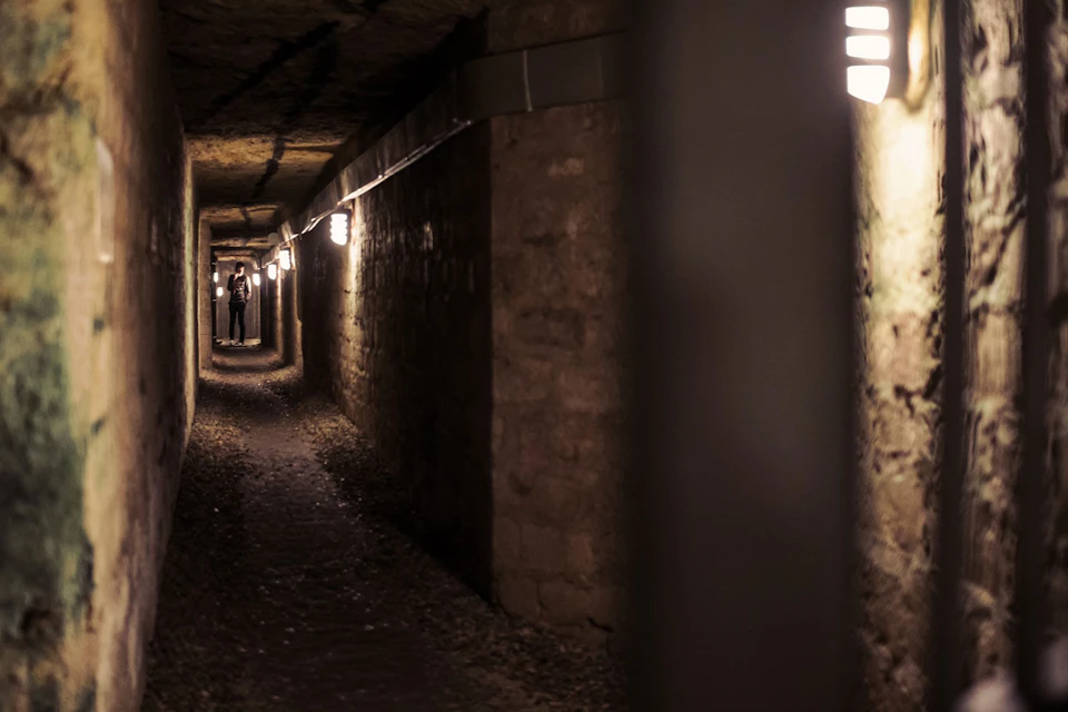 A dimly lit underground tunnel with rough walls and a person walking in the distance under electric lights.