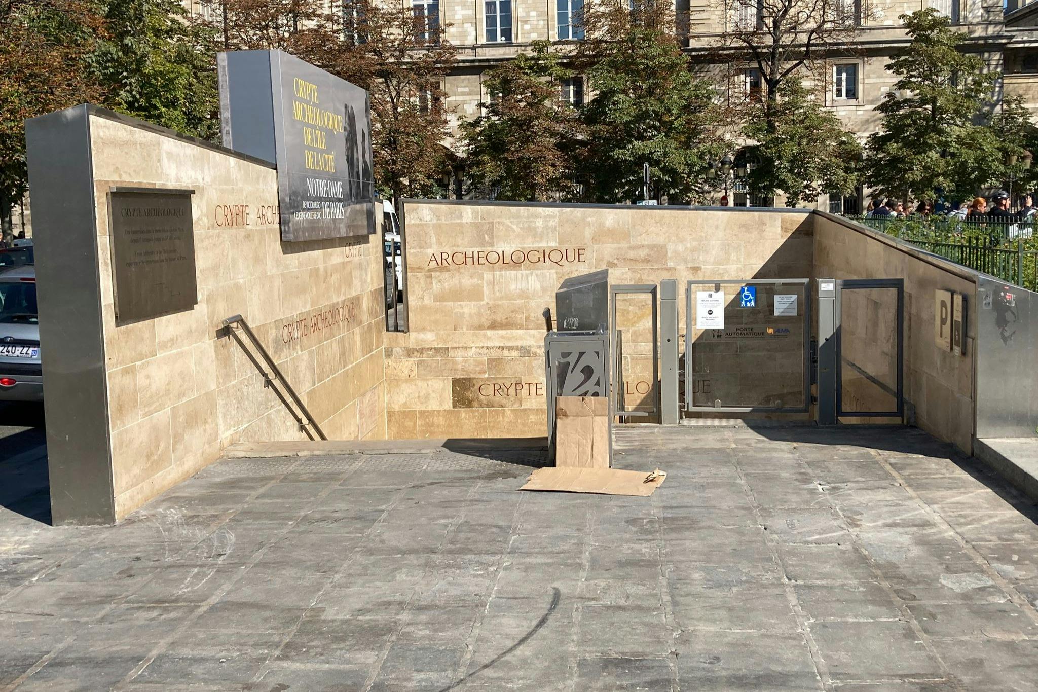 표지판과 계단이 있는 고고학 지하실의 돌 입구. 나무와 건물이 배경으로 보입니다.