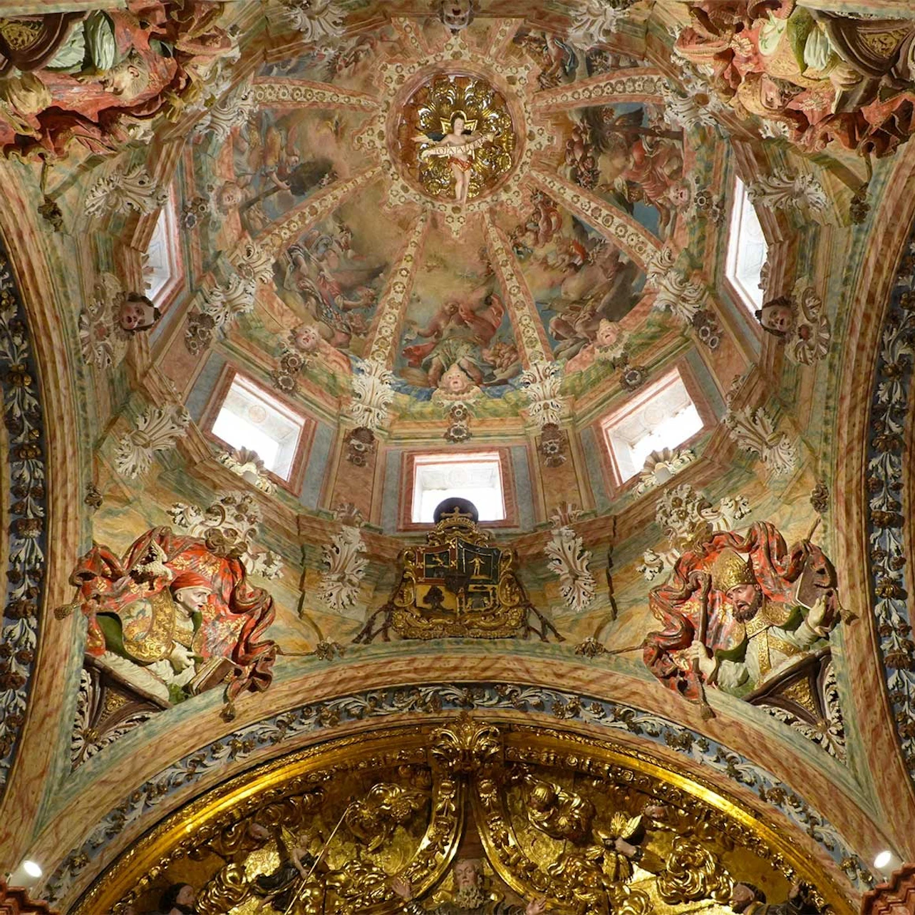 Ornate interior ceiling of a dome with intricate paintings, carvings, and golden decorative elements, illuminated by windows.