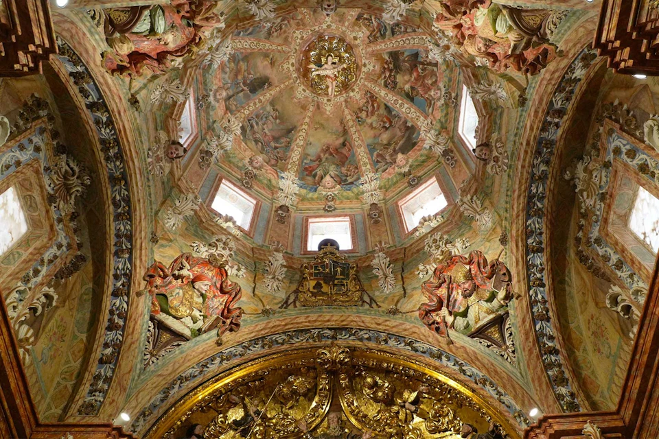 Ornate interior ceiling of a dome with intricate paintings, carvings, and golden decorative elements, illuminated by windows.