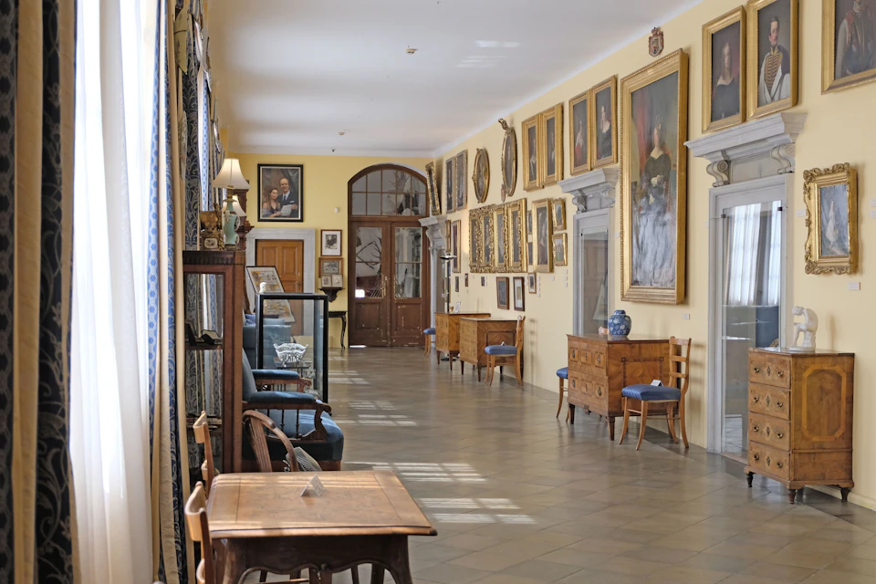 A hallway with framed paintings on the walls, wooden furniture, and display cabinets, lit by natural light through windows.