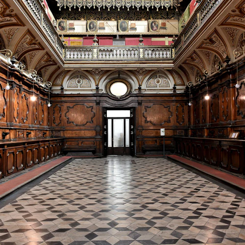 Ett stort, utsmyckat rum med träpaneler, invecklade tak, hängande lampor, rutigt golv och en stor frostad glasdörr.