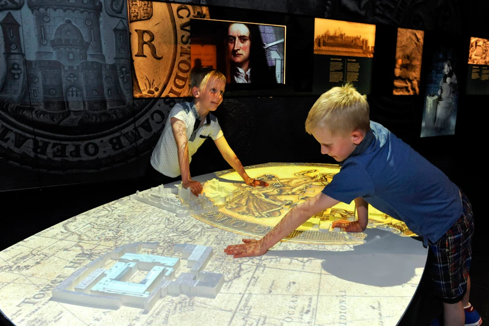 Two children interact with a large, illuminated map table in a museum exhibit featuring historical artwork and text panels on the walls.