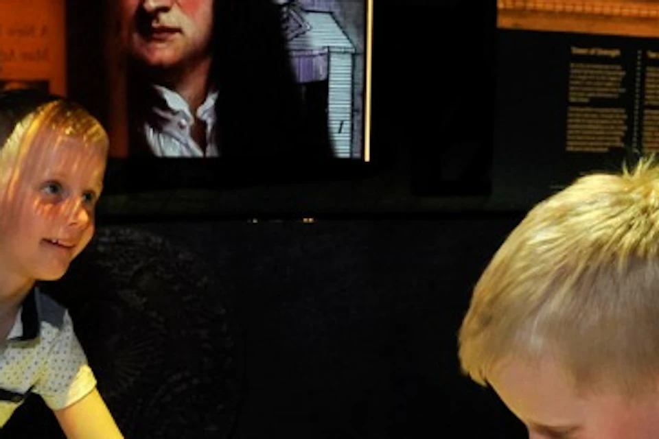 Two children interact with a large, illuminated map table in a museum exhibit featuring historical artwork and text panels on the walls.