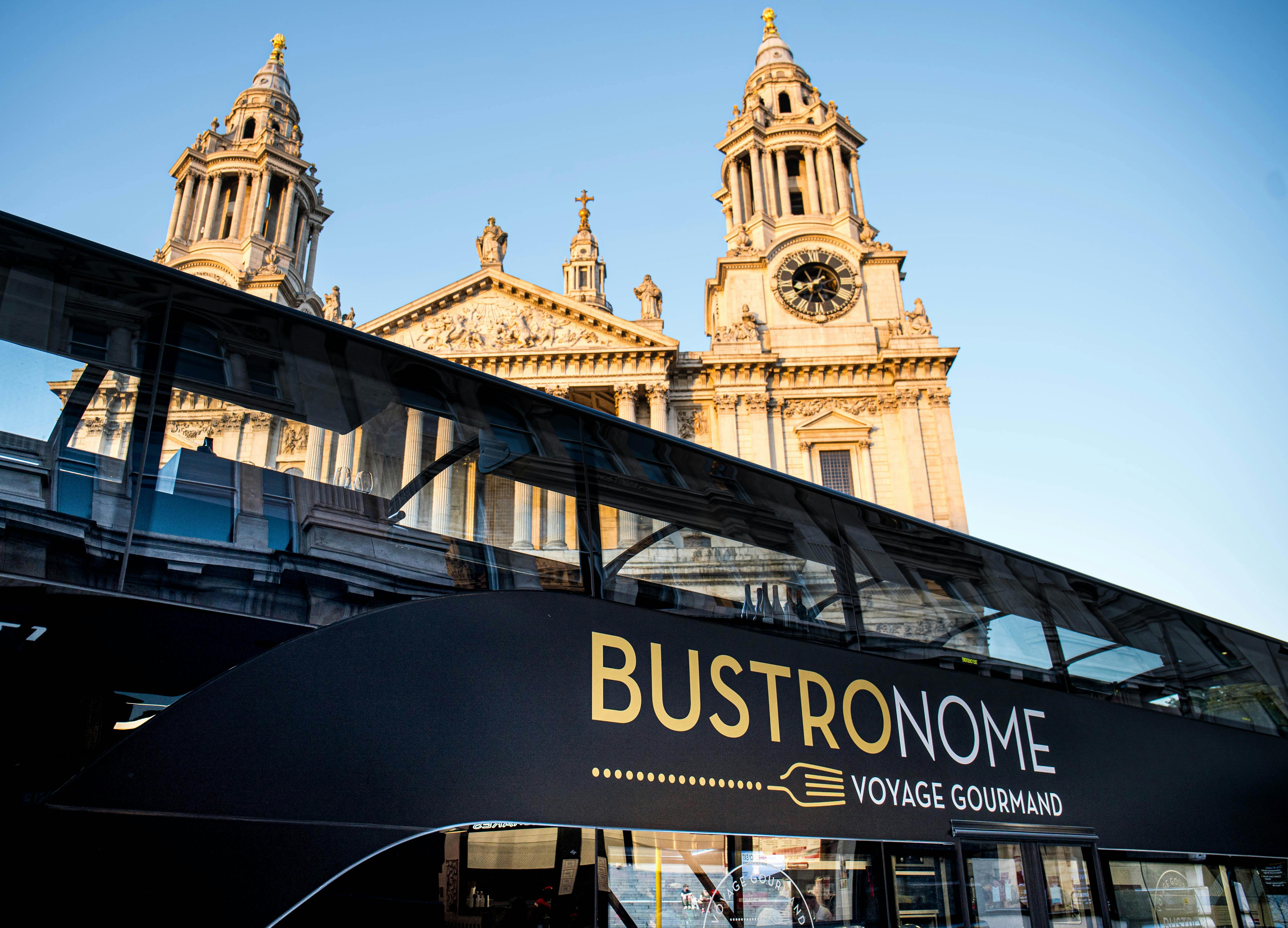 A tour bus with "Bustronome Voyage Gourmand" written on the side, parked near a historic church with clock towers.