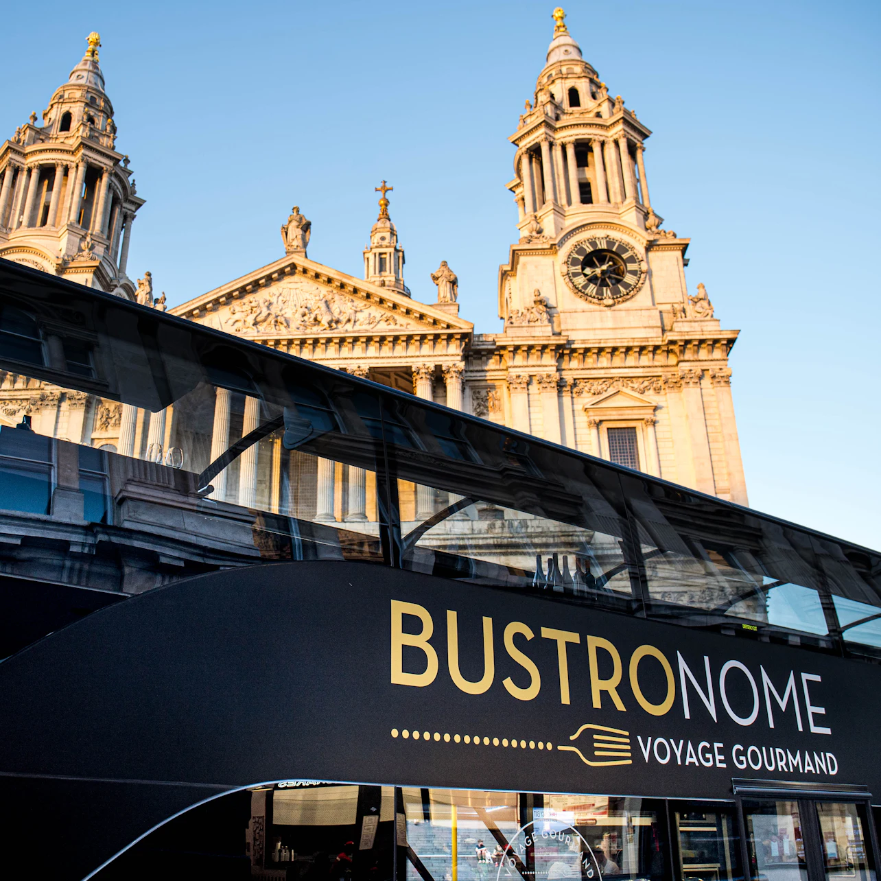 A tour bus with "Bustronome Voyage Gourmand" written on the side, parked near a historic church with clock towers.