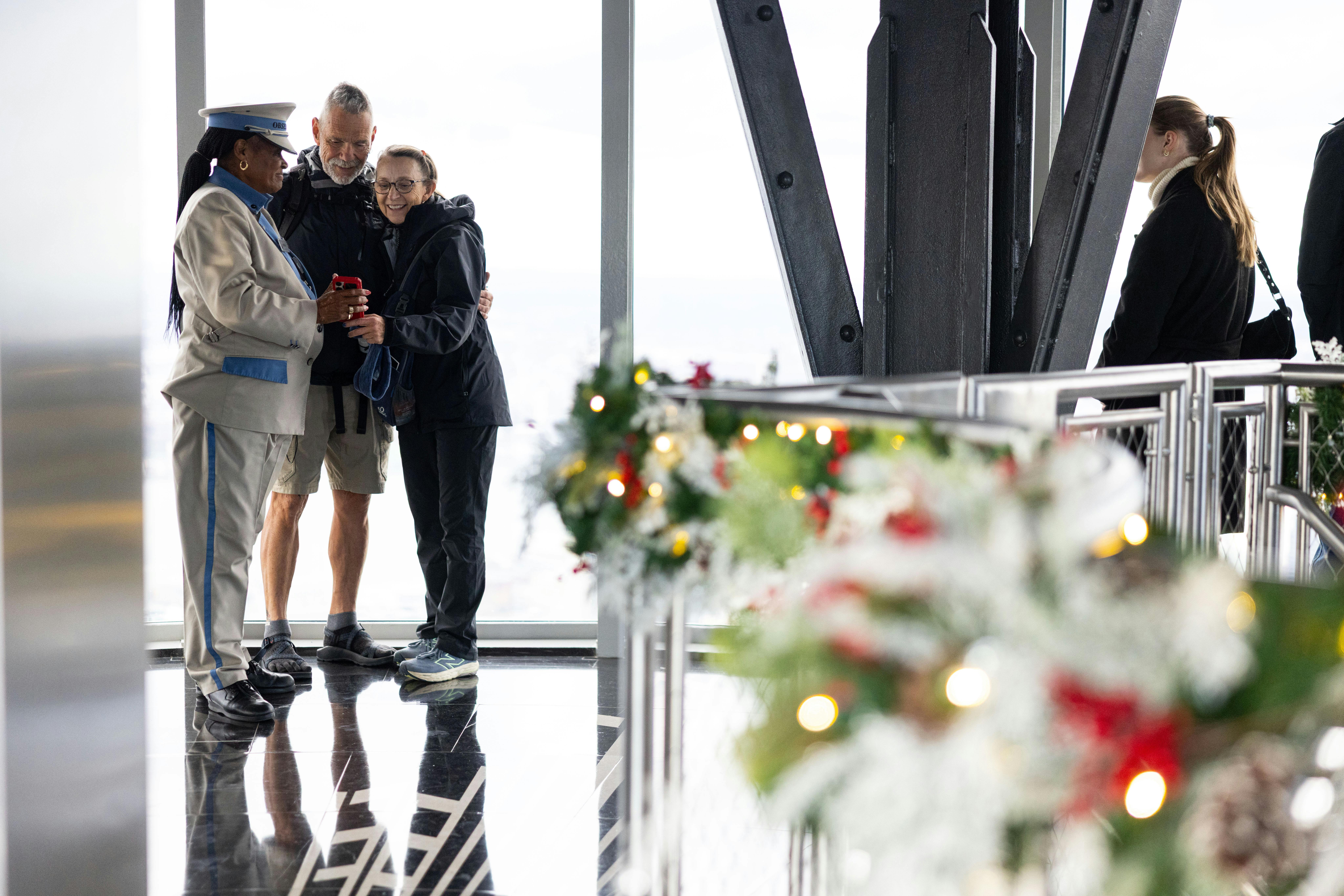 Ένας φρουρός ασφαλείας και δύο επισκέπτες στέκονται μαζί σε έναν σύγχρονο χώρο παρατήρησης με εορταστικές διακοσμήσεις ορατές σε πρώτο πλάνο.