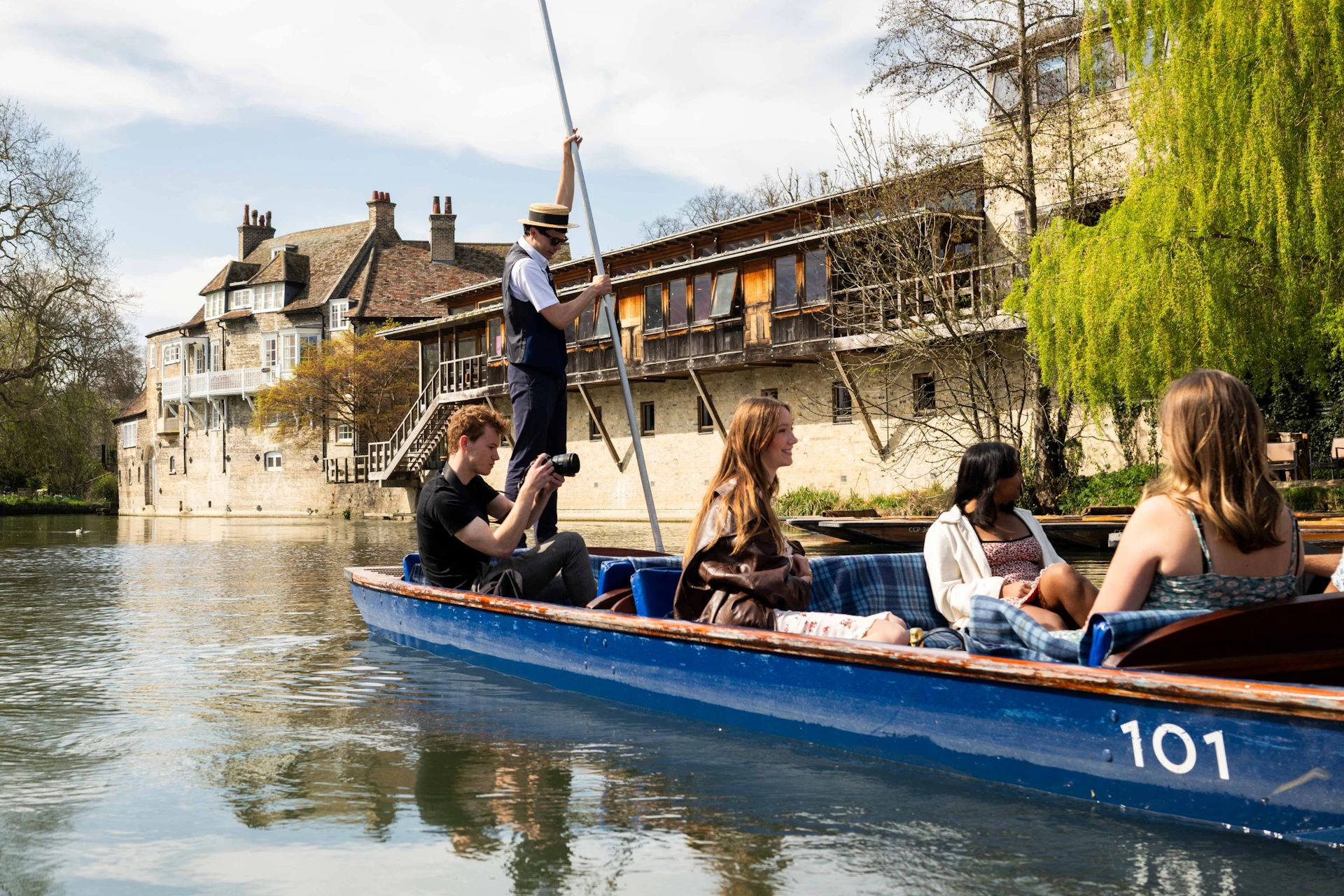 A group of people enjoying a punt ride on a river near traditional buildings and a leafy riverside under a sunny sky.