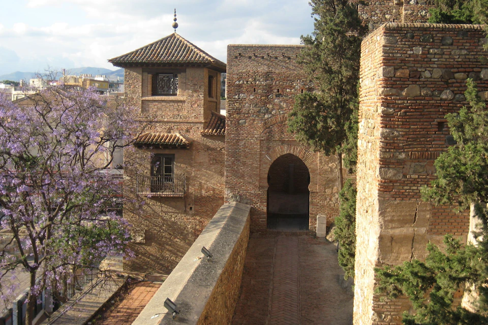 Historic stone building with arched doorway, tiled roofs, and tree on the right. Pathway leads towards the structure.