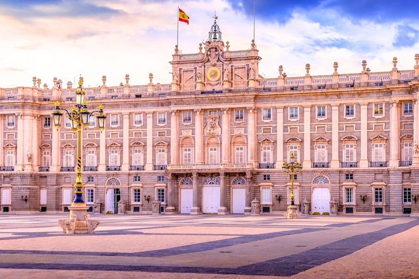 A grand palace with ornate architecture, a Spanish flag on top, and a spacious courtyard under a colorful sky.