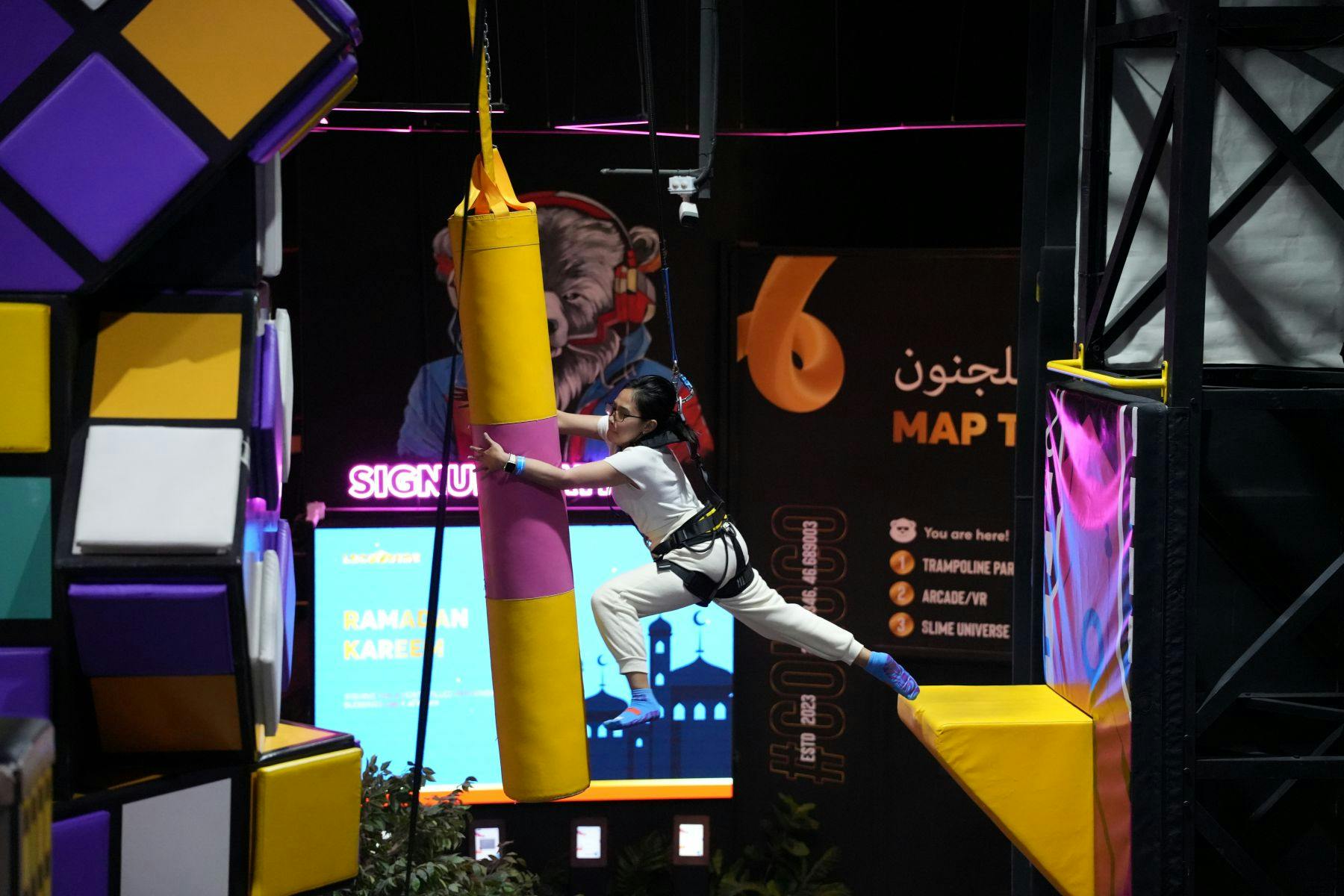 Ένα άτομο με ιμάντα πηδάει προς ένα αναρτημένο κίτρινο και ροζ παραγεμισμένο εμπόδιο σε μια εσωτερική εγκατάσταση αναψυχής.