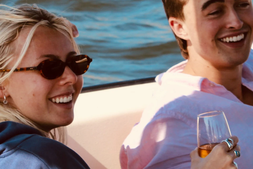 Two people sitting on a boat, smiling and holding glasses, with blue water and a bridge structure in the background.