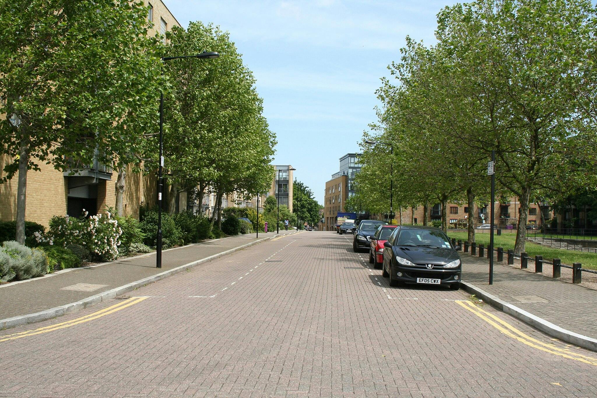 Une rue tranquille bordée de voitures garées et d'arbres, flanquée de bâtiments en briques et de verdure sous un ciel clair.