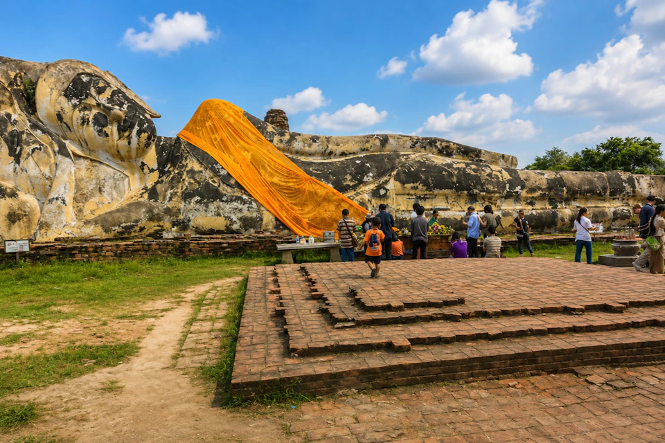 People gathered near a large reclining Buddha statue draped with an orange cloth against a blue sky with clouds.