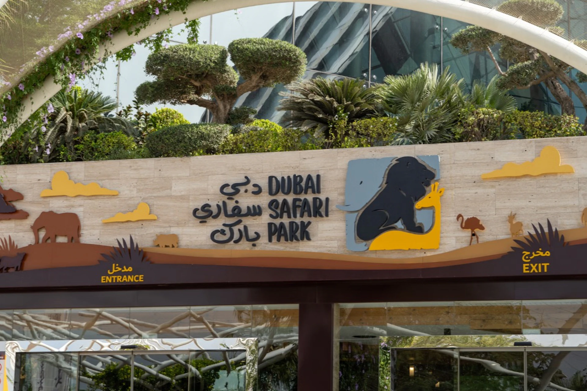 Entrance sign for Dubai Safari Park with text in English and Arabic, featuring a lion silhouette and surrounded by lush greenery.