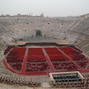 An empty ancient amphitheater with rows of red chairs arranged on the floor and a stage at the center, under a cloudy sky.