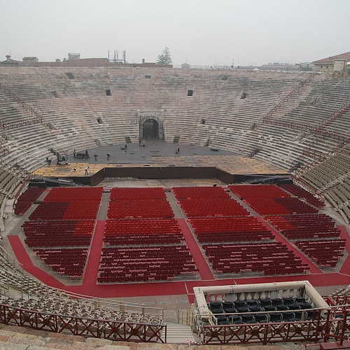 An empty ancient amphitheater with rows of red chairs arranged on the floor and a stage at the center, under a cloudy sky.