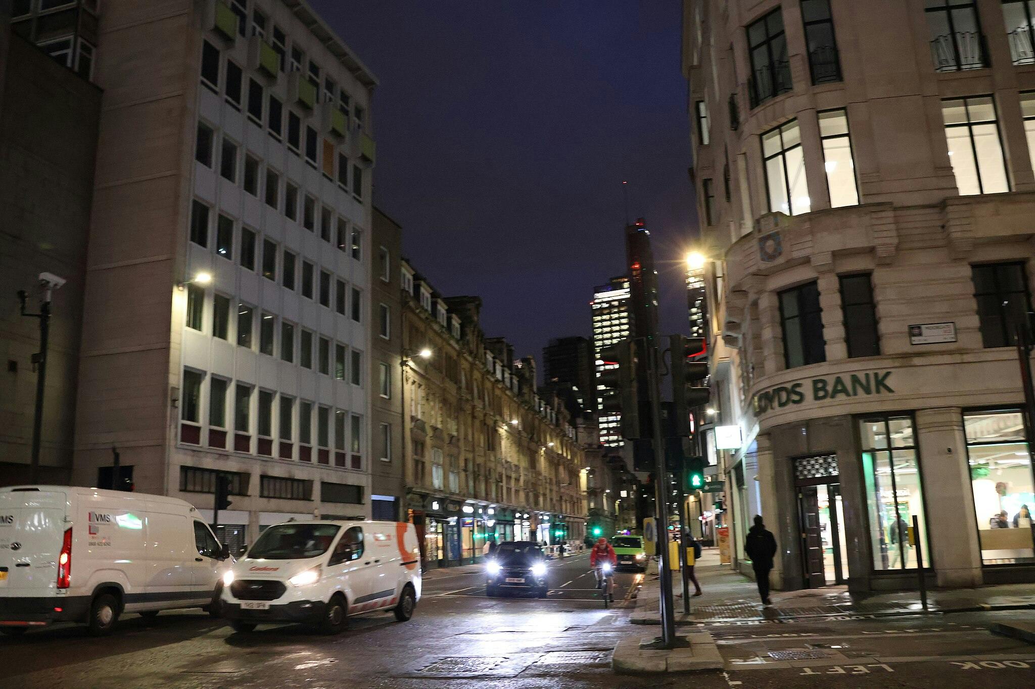 Eine nächtliche Stadtstraße mit beleuchteten Gebäuden, fahrenden Fahrzeugen, darunter Lieferwagen und ein Taxi, Radfahrern und Fußgängern.