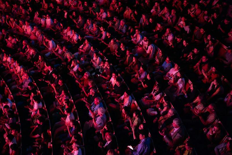 A packed theater audience seated in rows, illuminated by red lighting, facing a stage out of view.
