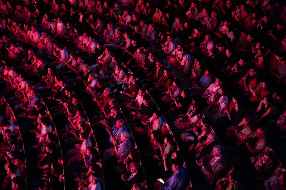 A packed theater audience seated in rows, illuminated by red lighting, facing a stage out of view.