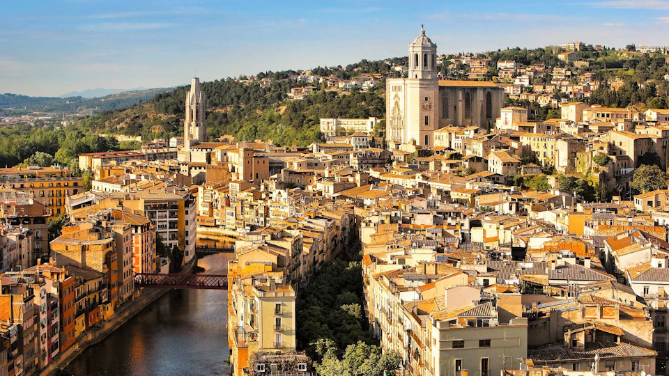 Aerial view of a sunlit city with historic buildings, a large cathedral, a river, and a background of green hills.