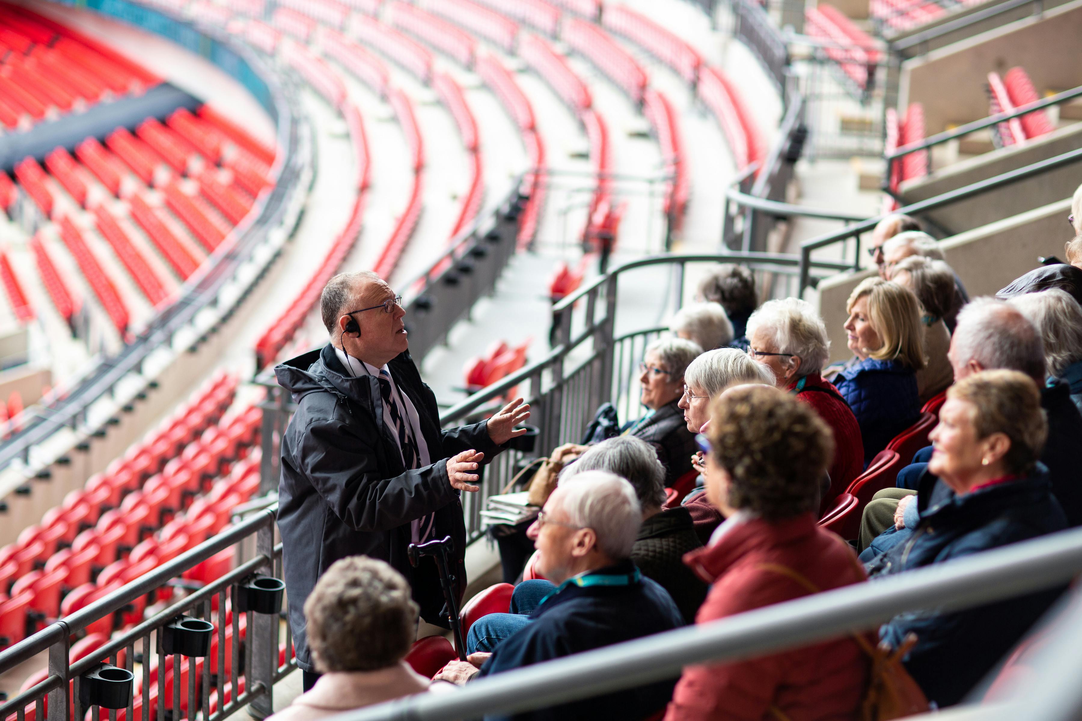 导游在红色座位和弧形排的体育场内与一群坐着的老人交谈。