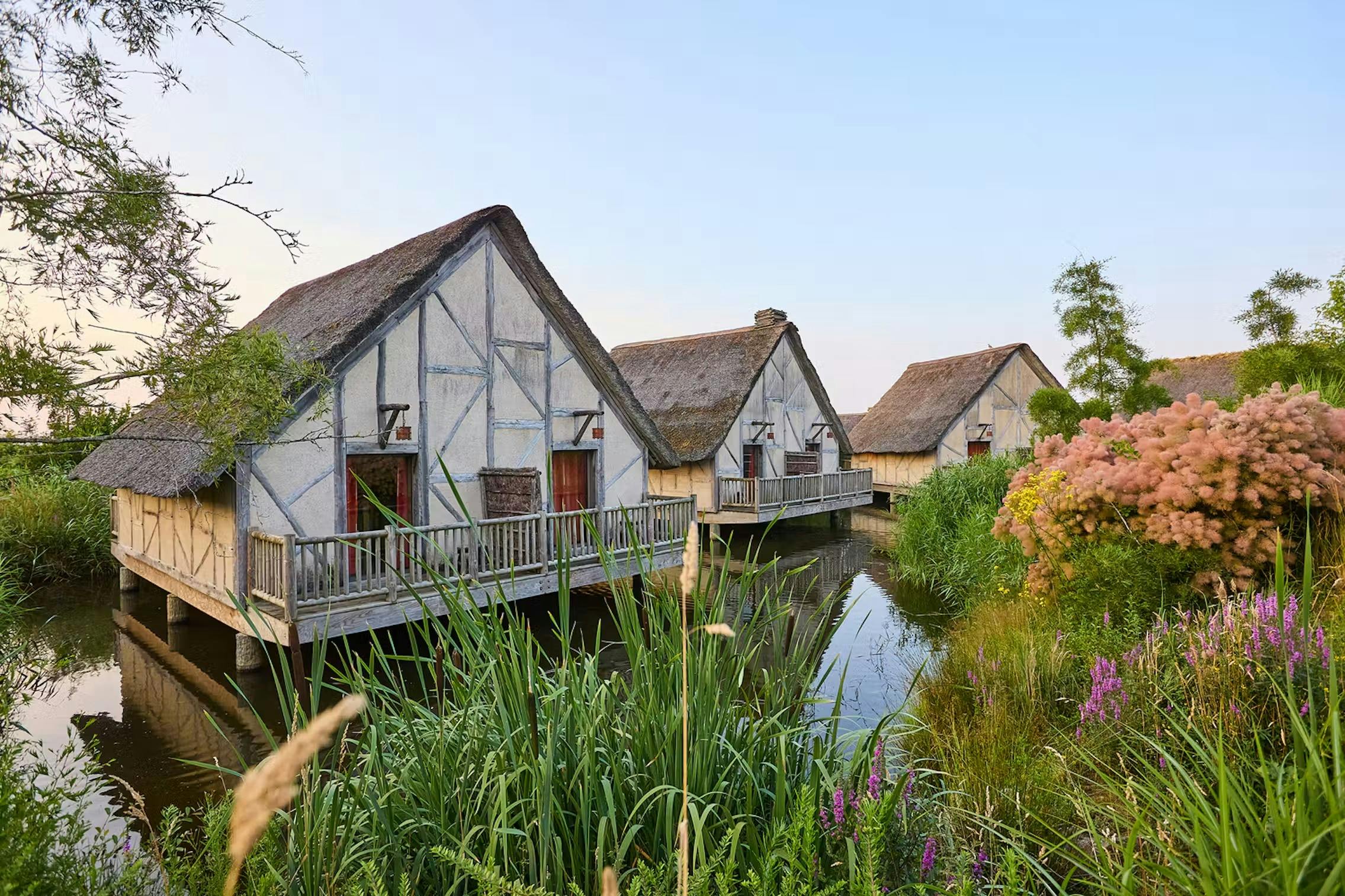 Three thatched cottages on stilts above a pond, surrounded by greenery and reeds, under a clear sky.