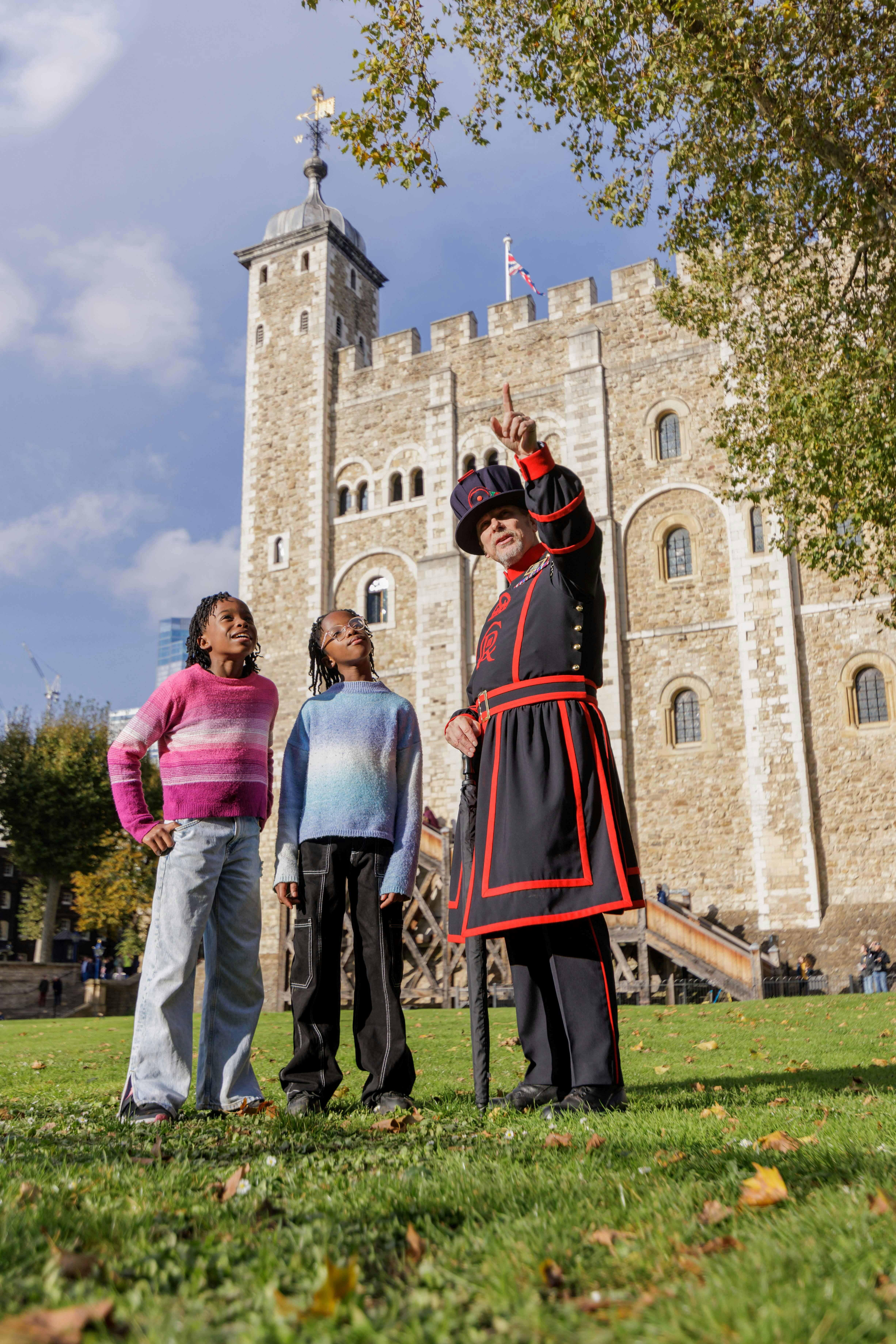 Yeoman Warder mluví se dvěma dětmi v barevných svetrech před Tower of London za slunečného dne.