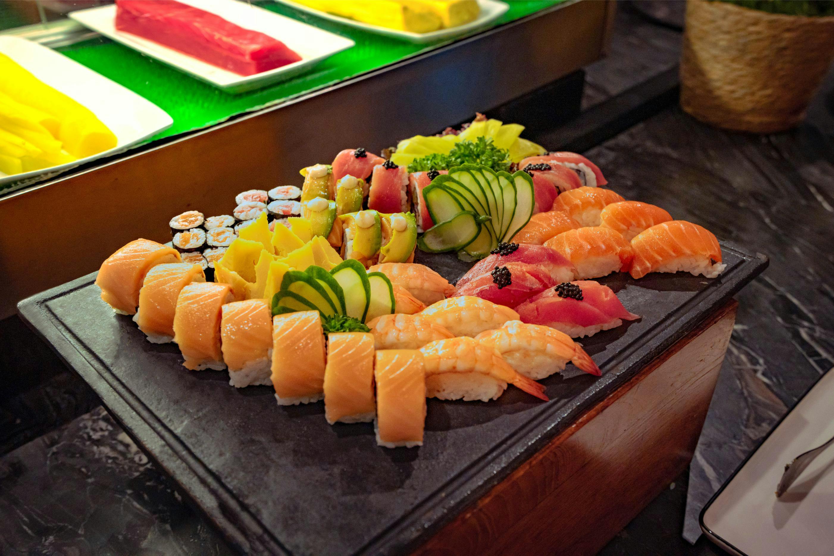 A platter of assorted sushi including nigiri and rolls, garnished with cucumber slices and herbs on a dark tray.