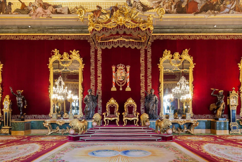 A lavish throne room with red drapery, golden thrones, ornate mirrors, chandeliers, and decorative sculptures.