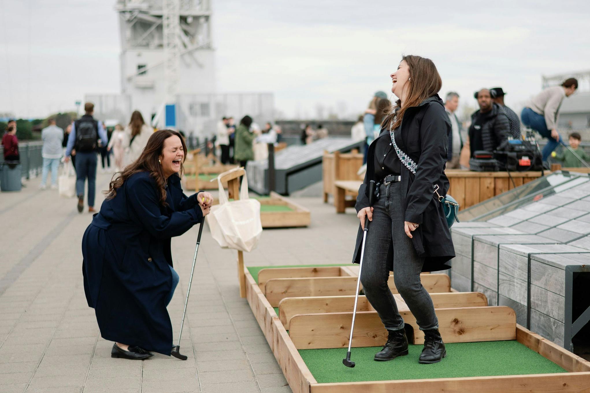 Duas mulheres riem enquanto jogam mini-golfe num campo exterior com barreiras de madeira. Outras pessoas são visíveis em segundo plano.