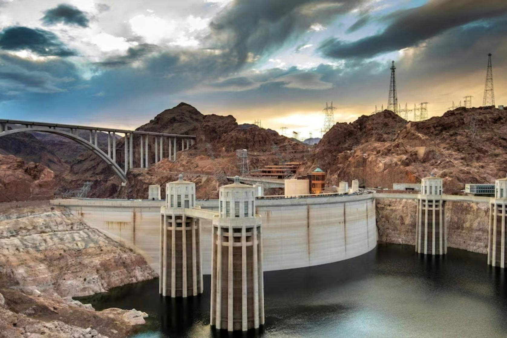 A dam with water towers on a river, surrounded by rocky hills, with a bridge and power lines in the background at sunset.