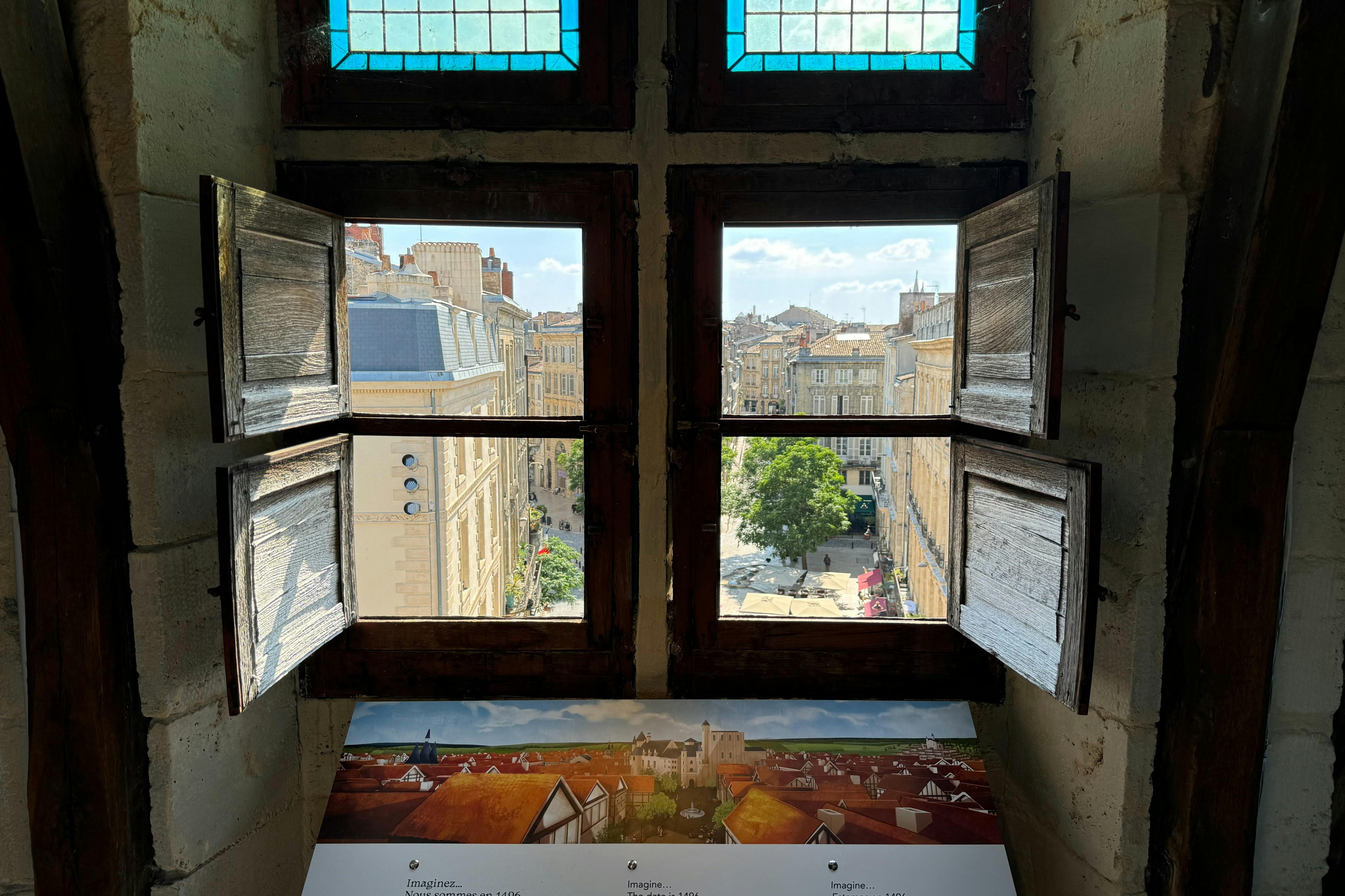 Open wooden shutters reveal a cityscape of historic buildings and streets under a blue sky with scattered clouds.